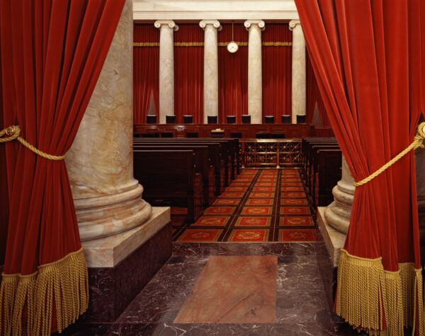 The Supreme Court chambers, showing the aisle between the spectator seats and the justices' seats at the end of the room. In the foreground are two marble pillars fronted with red velvet curtains with a heavy gold fringe at the bottom and gold tiebacks.