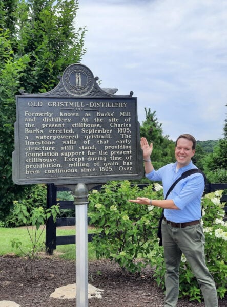 A white man in a blue shirt stands with his arms posed to present a historic marker next to him. The marker reads: "Old Gristmill-Distillery. Formerly known as Burks' Mill and distillery. At the site of the present stillhouse, Charles Burks erected, September 1805, a waterpowered gristmill. The limestone walls of that early structure still stand, providing foundation support for the present stillhouse. Except during time of prohibition, milling of grain has been continuous since 1805. Over"